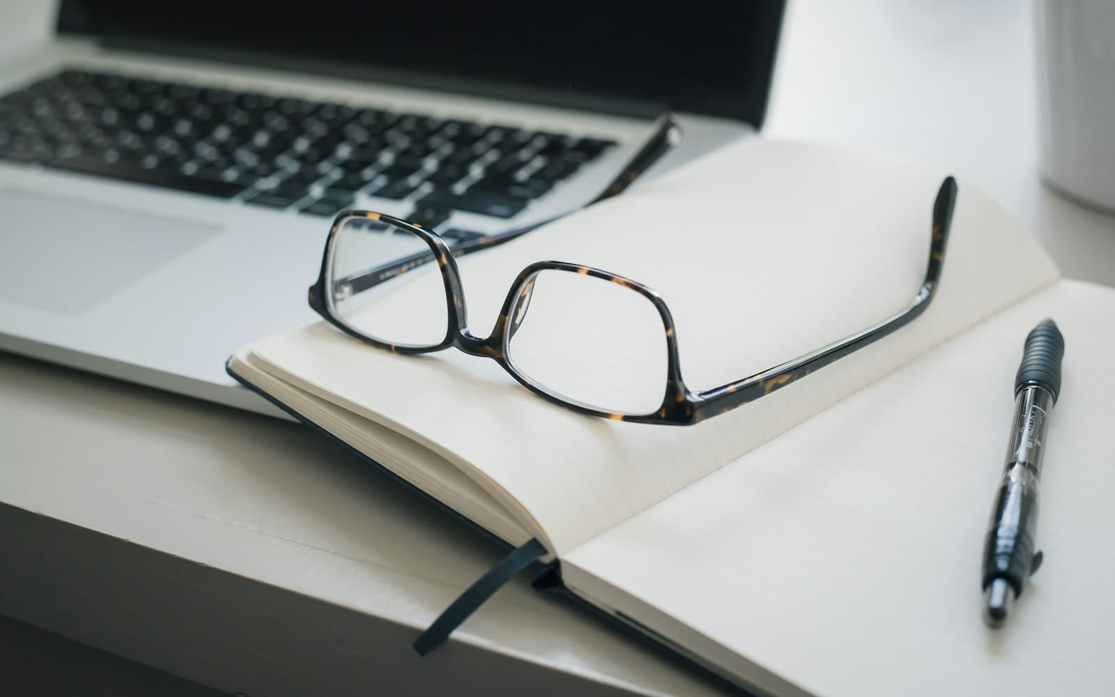 Reading glasses and a pen on an open notebook beside a laptop.