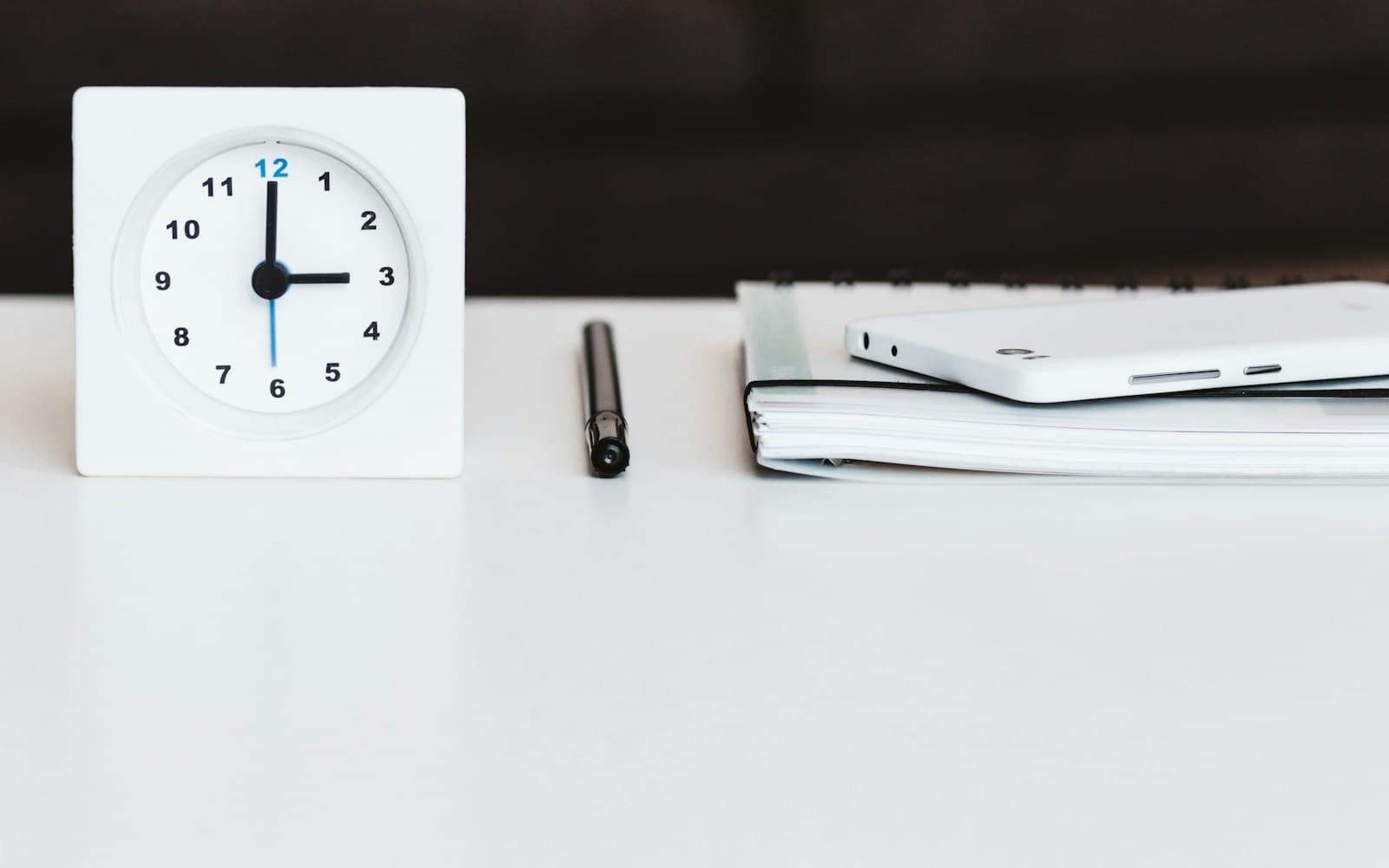 White analog clock beside a notebook, pen and mobile device.